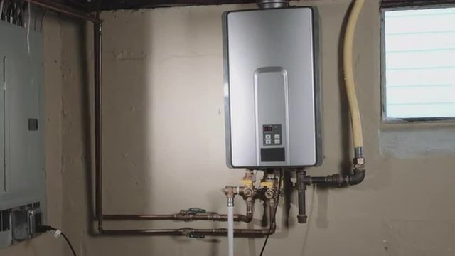 camera moves left from behind a wall to reveal a tankless water heater on the wall in a basement