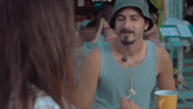 Slow motion of two friends sitting in outdoor cafe. Young man with beard wearing bucket hat listening to his smoking long-haired old chap