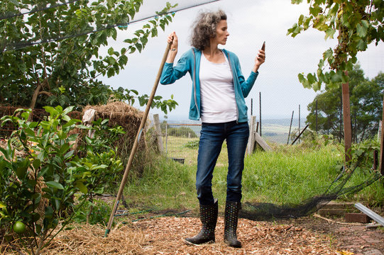Profile of mature woman in blue top and jeans in large garden, leaning on rake handle and using smart phone