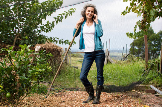 Mature woman with grey hair and jeans in large garden, leaning on rake handle and smiling while using smart phone