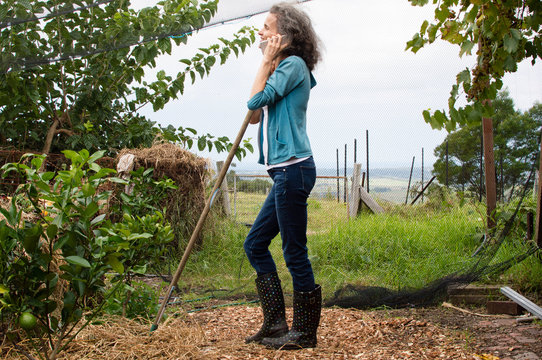 Profile of mature woman in blue jeans leaning on rake handle in garden and using smart phone
