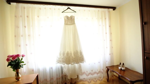 Close up of a fashionable wedding dress on a hanger , white shoes on the floor and a bouquet with pink roses in the room of the bride.