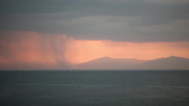 Timelapse shot of of heavy cloudy with rain pouring sailing over the sea and distant coastal city, lightning strikes and and then twilight coming