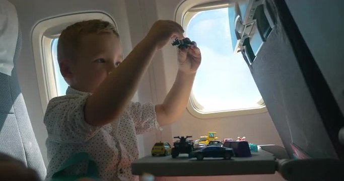 Little boy traveling by air. He sitting by the illuminator and playing with toy plane, many cars on the table in front of him. Entertaining during the flight
