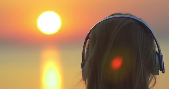 Close-up back shot of a blond woman putting on wireless headphones, golden sunset over sea as background. Relaxing outdoor with favourite music