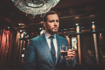 Confident well-dressed man with glass of whisky in luxury apartment interior.