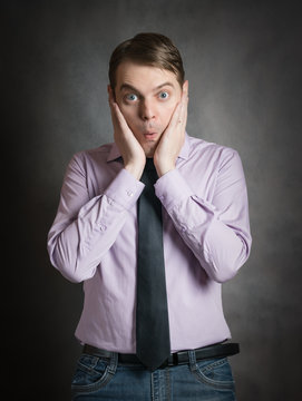 Portrait of pensive young man in pink shirt and dark tie with surprised face, against dark background.