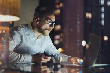Bearded young businessman working on modern loft office at night. Man using contemporary notebook texting message, holding smartphone, bokeh background. Horizontal, film effect. 