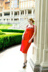 Obraz premium Attractive young woman in red dress posing on stairs of luxurious house with bright green garden on background