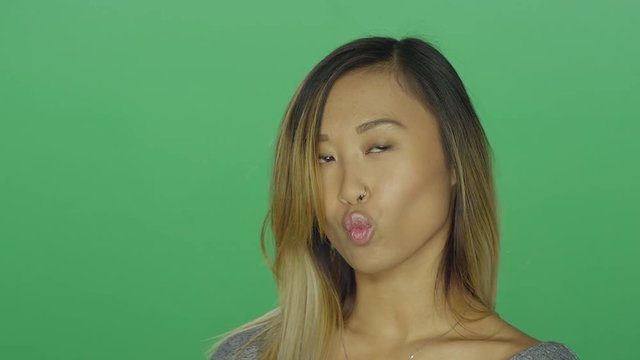 Cute Asian girl making a kissy face and smiling, on a green screen studio background