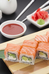 Salmon sushi roll on wooden plate ( gete ) with ginger wasabi chopsticks and sauces over black background