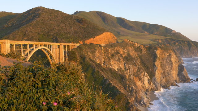 Locked down and panning view of waves breaking on the coastline at Pacific Coast Highway's Bixby Bridge at sunset in the Big Sur area of Central California