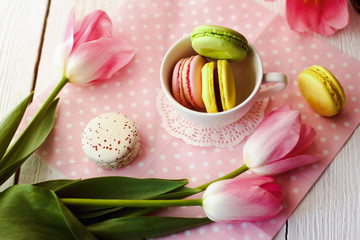 Obraz na płótnie Canvas A beautiful flowers pink tulips with colorful macaroons laid in cup on white wooden background with pink Lacy napkin