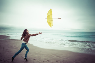 Un jeune  femme tentant de rattraper son parapluie jaune emporté par le vent sur une plage 