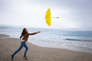 Fototapeta na wymiar Un jeune femme tentant de rattraper son parapluie jaune envolé sur une plage 