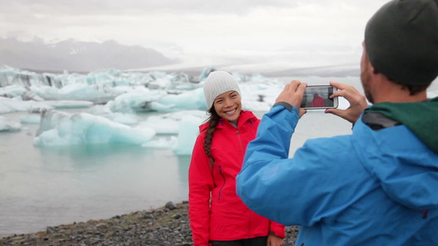 Iceland - couple taking photo with smartphone by Jokulsarlon glacial lagoon / glacier lake. Happy tourists on travel enjoying beautiful Icelandic nature landscape with Vatnajokull in background.