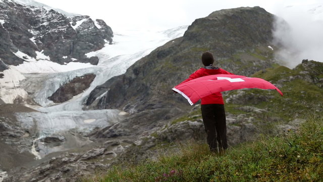 Switzerland hiker hiking cheering showing Swiss flag jumping in front of glacier. Happy Asian woman holding big red flag in nature.
