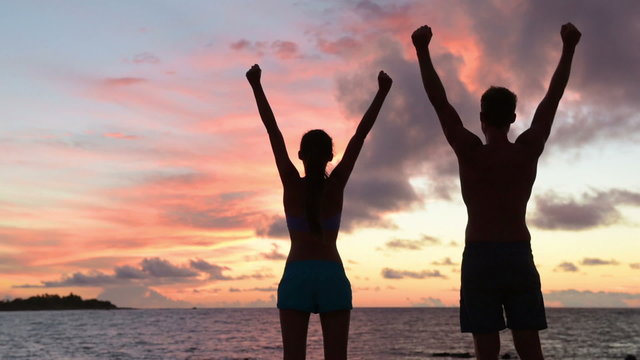 Success, winning and achievement and accomplishment concept with fitness people cheering and celebrating of joy with arms raised outstretched up by ocean beach outdoors. Fitness couple at sunset.