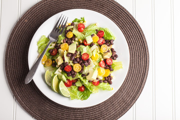 Top view of homemade southwest black bean lime salad with corn, cherry tomatoes, lettuce, avocado, and black beans with vinaigrette dressing for clean eating on brown place mat 