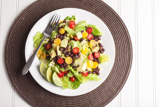 Top View Of Homemade Southwest Black Bean Lime Salad With Corn, Cherry Tomatoes, Lettuce, Avocado, And Black Beans With Vinaigrette Dressing For Clean Eating On Brown Place Mat 