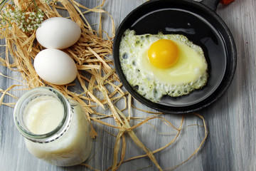 Overhead view of white eggs in egg cups surrounded by straw,  milk and a fried egg in an iron skillet on a wooden background with copy space.