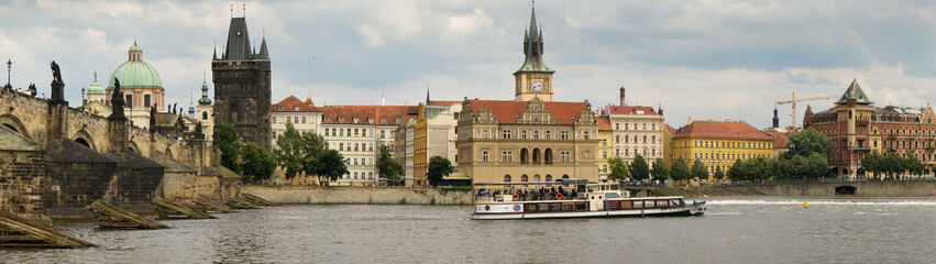 Fototapeta na wymiar Wide panorama looking towards the eastern bank of Prague’s Vlatava river. Charles Bridge, Saint Francis of Assisi Church, Old Town Bridge Tower, an historic water tower and a river cruise boat.