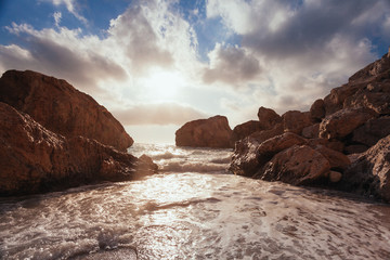 Beautiful beach landscape. Wave splashing on shore rocks. Colorful sunset in the background.