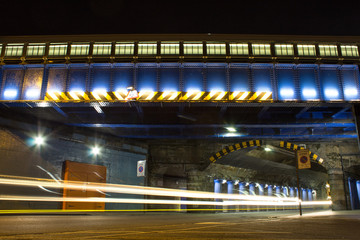 Train goes over the bridge at night with blurry lights and cars on the street
