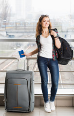 dark-haired young woman standing with a suitcase and a backpack and holding a passport with a ticket. Full length.