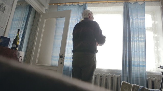 Senior opens the curtains in the room and the light gets in the room. Old grandfather with flowers mimosa came to greet his wife that Hvar lying on the bed.