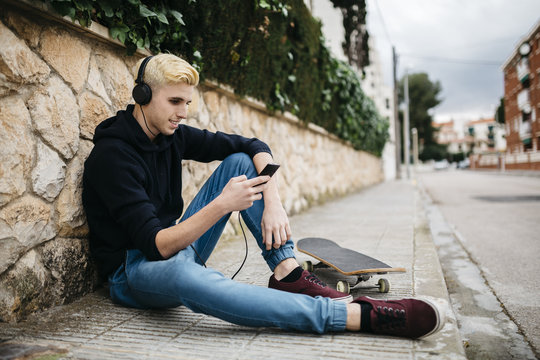Spain, Torredembarra, young skateboarder sitting on pavement listening music with headphones