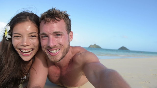 Couple relaxing on beach taking selfie picture with camera smartphone. Young multiracial couple on getaway vacation in Hawaii lying down looking at camera. Candid closeup angle looking real.