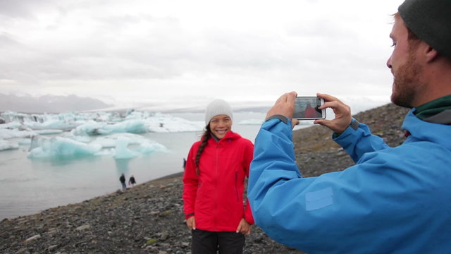 Couple taking photo with smartphone by Jokulsarlon glacial lagoon / glacier lake on Iceland. Happy tourists on travel enjoying beautiful Icelandic nature landscape with Vatnajokull in background.