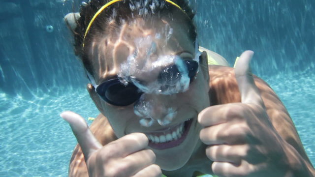 Woman swimming underwater in pool smiling happy giving thumbs up sign hand waving hands saying hello looking at camera. Young female swimmer with swim goggles at holiday resort.
