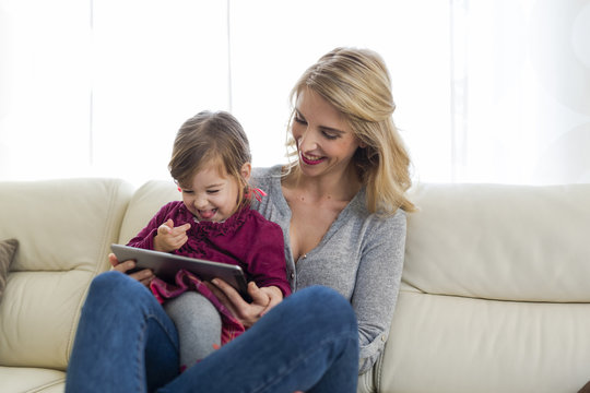 Mother and her little daughter sitting on couch in the living room looking at digital tablet