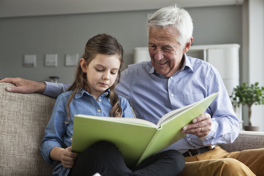 Grandfather and his granddaughter sitting together on the couch with a book