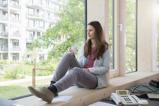 Portrait of creative business woman sitting on window sill having coffee break