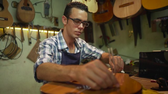 Lute maker shop and classical music instruments: young adult artisan fixing old classic guitar, tuning the instrument with a metallic diapason. Closeup of hand on guitar body