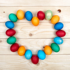 Fototapeta na wymiar Heart-shaped frame of colored Easter eggs over the light wooden surface as copyspace festive background composition