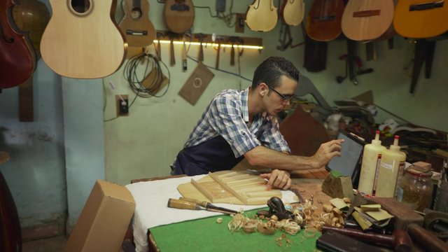 Lute maker shop and acoustic music instruments: artisan cutting wood to make a classic guitar. He reviews the project on tablet pc. Closeup of his hand measuring the guitar body. Steadicam shot