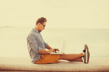 Young man working on laptop while sitting on the beach on the pier, sending mail, mounts, video editing and photography,working abroad,freelance working,typing online,social networks,periscope,wi-