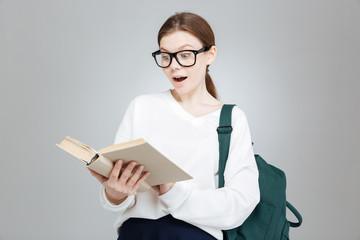 Surprised cute girl student in glasses with backpack reading book