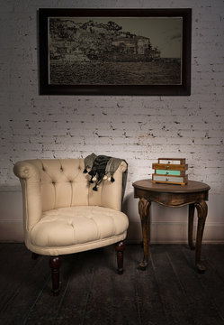 Vintage still life of old beige armchair, small drawers unit, hanged framed painting and one small antique table on dark brown wooden floor and white bricks wall in Studio