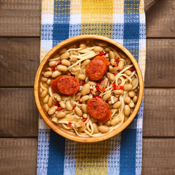 Traditional Chilean dish called Porotos con Riendas (English: beans with reins), made of cooked beans, linguine (flat spaghetti) and served with fried sausage, photographed on wood with natural light