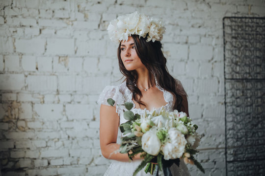 girl with a wreath of white flowers on her head in a white wedding dress holding a bouquet of white flowers and greenery on the background of white brick wall