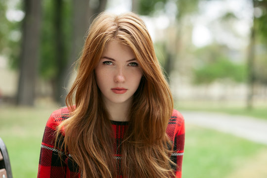 Closeup portrait of young adorable redhead woman in red plaid jacket stares into camera with blurred park background