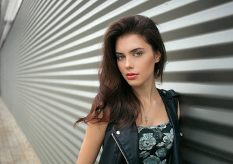 Closeup portrait of young beautiful brunette woman in black leather jacket posing outdoors against urban style background of metal strips