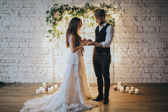 girl in a white wedding dress and bearded man in suit and hat standing near the ceremonial arches of lights and green branches on a background of white brick wall