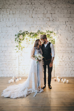 girl in a white wedding dress and bearded man in suit and hat standing near the ceremonial arches of lights and green branches on a background of white brick wall