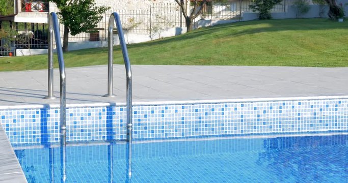Cinemagraph - outdoor pool with stairs on resort or house area. Clear blue water rippling in shining in sunlight, green lawn in background. Recreation time
